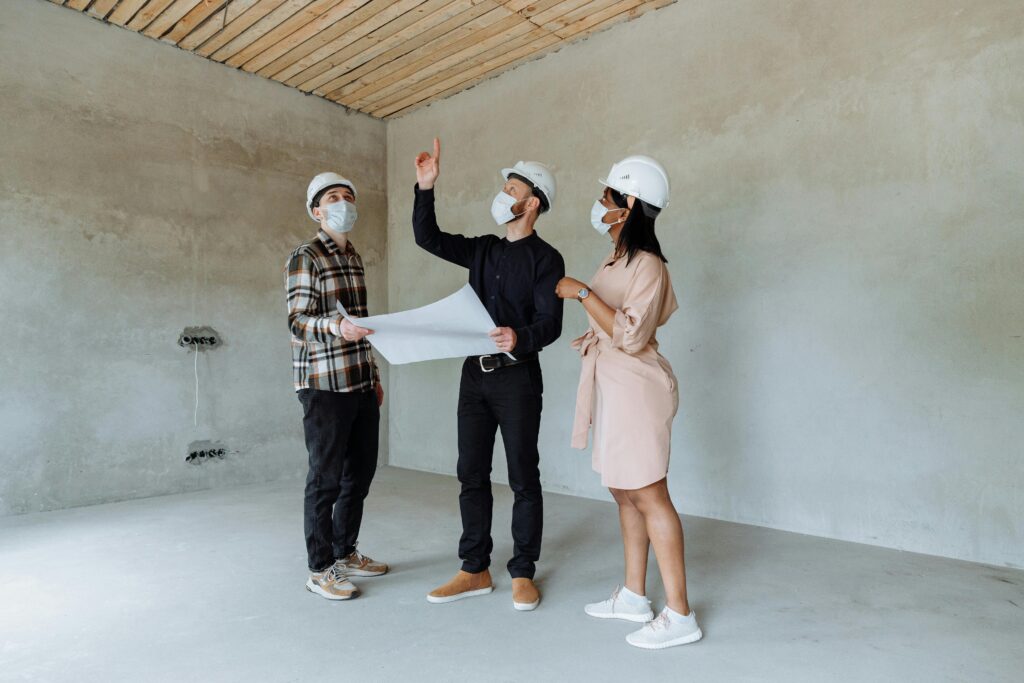 Three architects wearing face masks and helmets discuss plans in an indoor construction site.